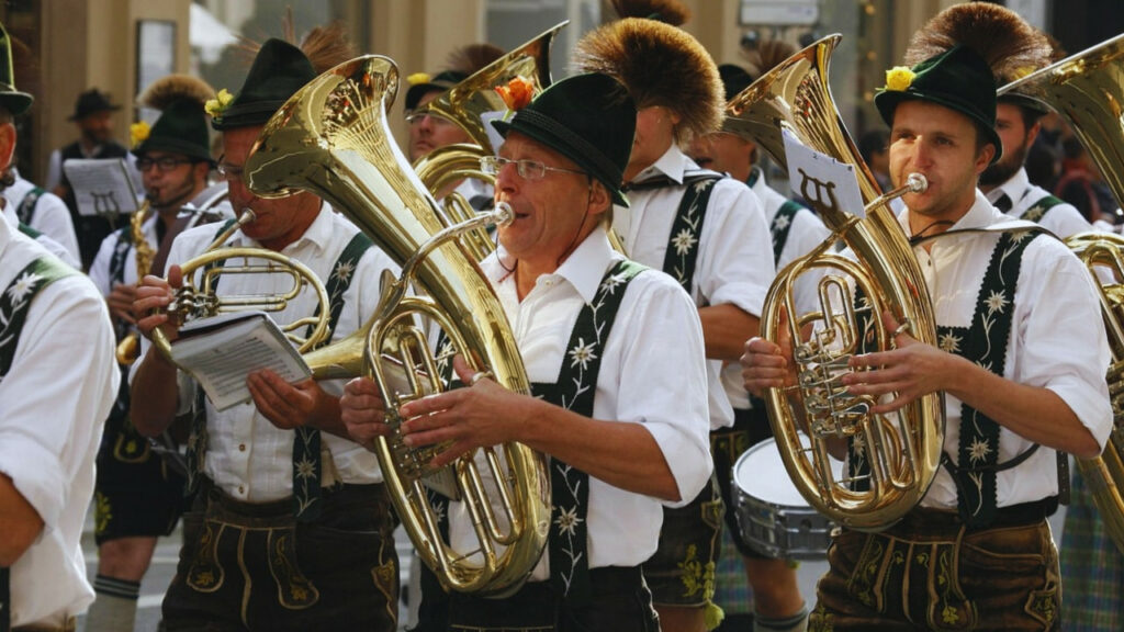Grupo de músicos da Oktoberfest tocando instrumentos de sopro durante desfile tradicional, com trajes típicos germânicos.